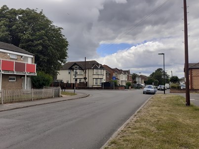 A photo of Radstock Road at the junction of Manor Road South before the works have taken place. The road and nearside grass verge are in the foreground, with housing to the left, centre background and right. The road is mostly clear, except for some parked cars to the right of the photo.