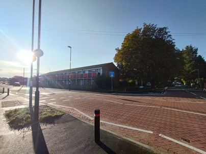 A photo of Radstock Road at the junction with Manor Road South after the works have been completed. The road itself has a new red paved surface, with new bollards on the nearside pavement and a new zebra crossing. The sun is rising behind the nearside zebra crossing light.