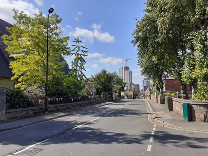 A photo of Weston Grove Road prior to the works being undertaken. St Mark's Church is on the left, partially obscured by trees. Trees within the boundary of Woolston Community Centre are to the right, and in the distance can be seen Centenary Quay.